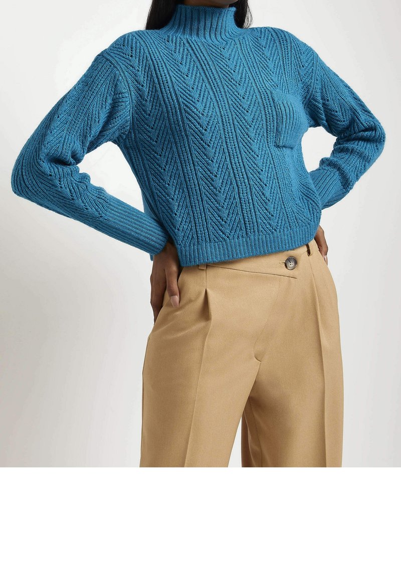 Maglione blu lavorato a maglia con un collo alto, caratterizzato da un motivo a spina di pesce strutturato. Abbinato a pantaloni beige chiaro, taglio sartoriale, con dettaglio dei bottoni.