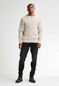 Jasnobeżowy sweter z dzianiny z wzorem warkocza, okrągłym dekoltem i ściągaczami przy mankietach, połączony z ciemnymi dżinsami i brązowymi butami.