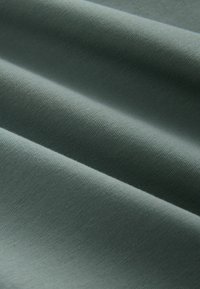Gros plan d’un tissu doux, lisse, gris clair avec une texture fine visible et des plis doux créant des lignes d’ombre subtiles.
