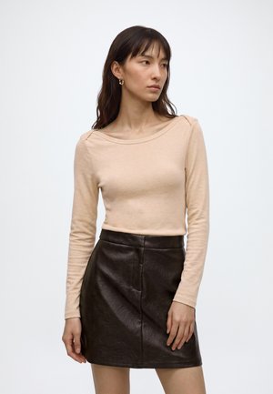 Jeune femme aux longs cheveux foncés, portant un haut beige à manches longues et une jupe en cuir noire, debout devant un fond blanc uni.
