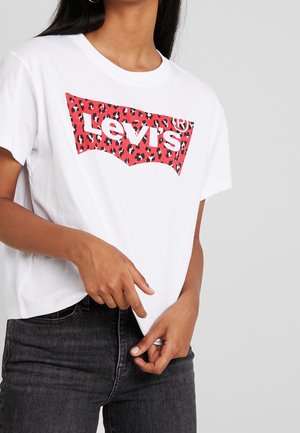 Femme portant un t-shirt blanc Levi's avec un logo imprimé léopard rouge, associé à un jean gris foncé, sur un fond uni.