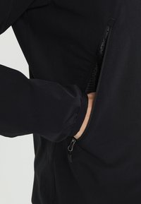 Veste noire au design élégant avec un tissu texturé, dotée d'une poche zippée sur le côté et de poignets élastiqués.