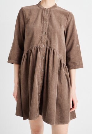 Robe en velours côtelé marron avec boutonnage sur le devant, taille froncée et manches trois-quarts portée par une personne sur un fond uni.