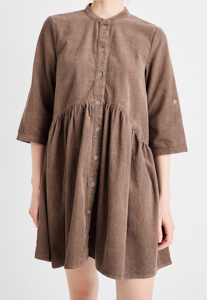 Robe en velours côtelé marron avec boutonnage sur le devant, taille froncée et manches trois-quarts portée par une personne sur un fond uni.