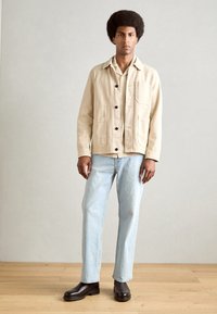 Beige cotton jacket with four pockets, black buttons, worn over a light shirt. Light blue jeans and black ankle boots complete the outfit.