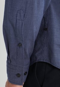 Niebieska koszula z długim rękawem w biały kratkowany wzór, z zapięciem na guzik na mankietach, noszona na czarnych spodniach, ukazująca szczegóły na bokach i rękawach.