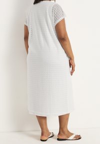 Kvinna klädd i en vit, texturerad knälång klänning och vita sandaler, stående mot en enkel ljus bakgrund med ryggen vänd mot kameran.