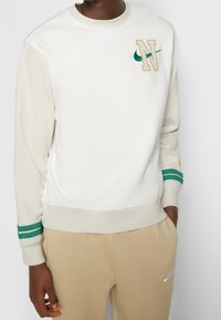 Vit och beige Nike-hoodie med gröna och guldiga detaljer, prydd med en broderad 'N' och en grön swoosh, ribbade ärmslut och en texturerad yta.