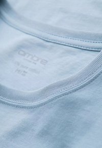 Světlomodré bavlněné tričko zblízka, s kulatým výstřihem a jemně žebrovanými okraji. Měkká textura s potiskem značky a údaji o údržbě uvnitř.