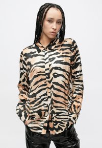 Satiinist pika varrukaga särk, millel on beeži ja musta tiigri triibumuster, nööpidega esiosa ja krae. Kangal on sile tekstuur.