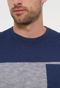 Άνδρας με κοντό μούσι φοράει μπλε σκουρό μπλουζάκι με οριζόντιο ανοιχτό γκρι και λευκό ριγέ μοτίβο στο στήθος.