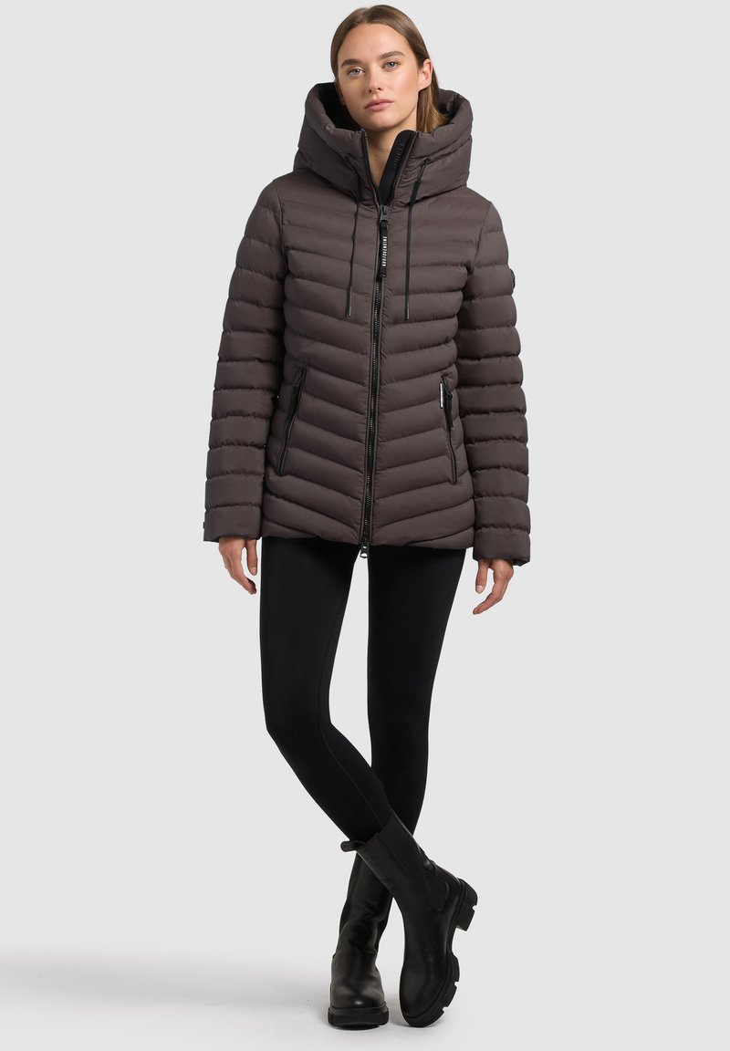Gepolsterte, kapuzenartige Jacke in Dunkelbraun mit diagonaler Steppung, Reißverschluss vorne und Seitentaschen. Getragen mit schwarzen Leggings und Stiefeln.