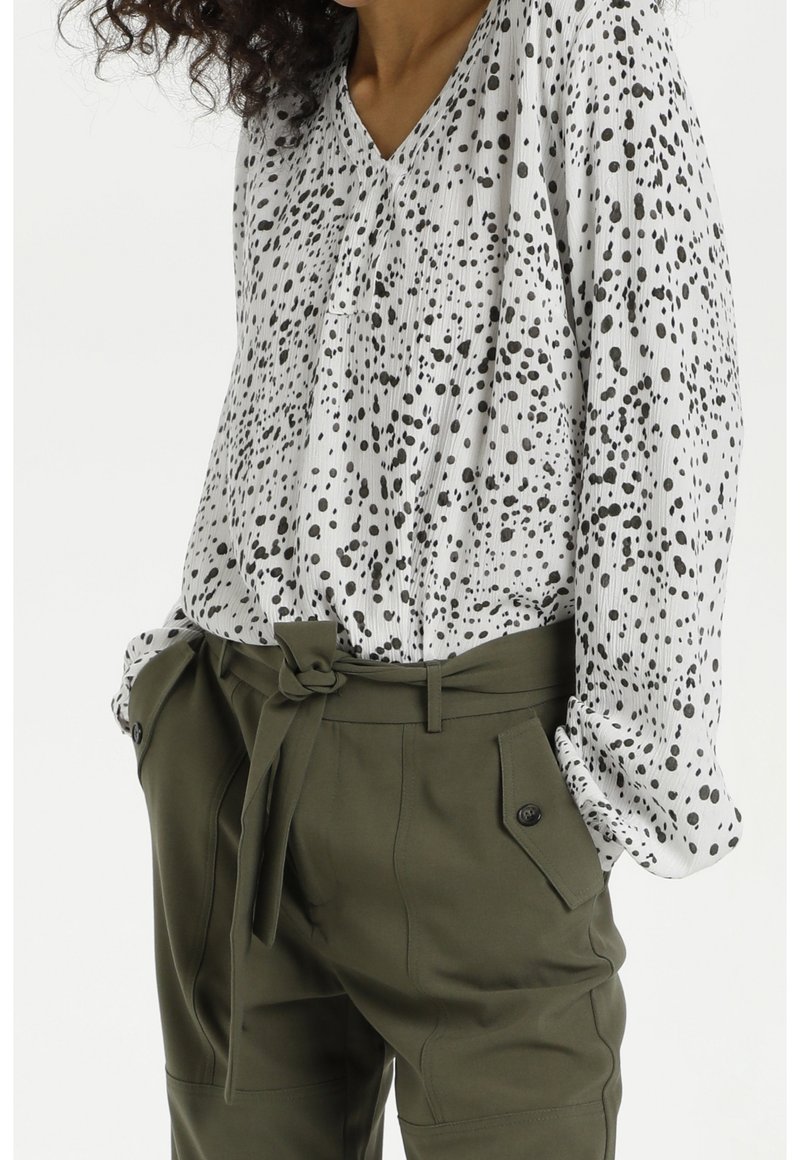 Vrouw draagt een witte blouse met lange mouwen met zwarte vlekken, ingestopt in olijfgroene, taillehoge broek met een strikceintuur en zakken met knopen.