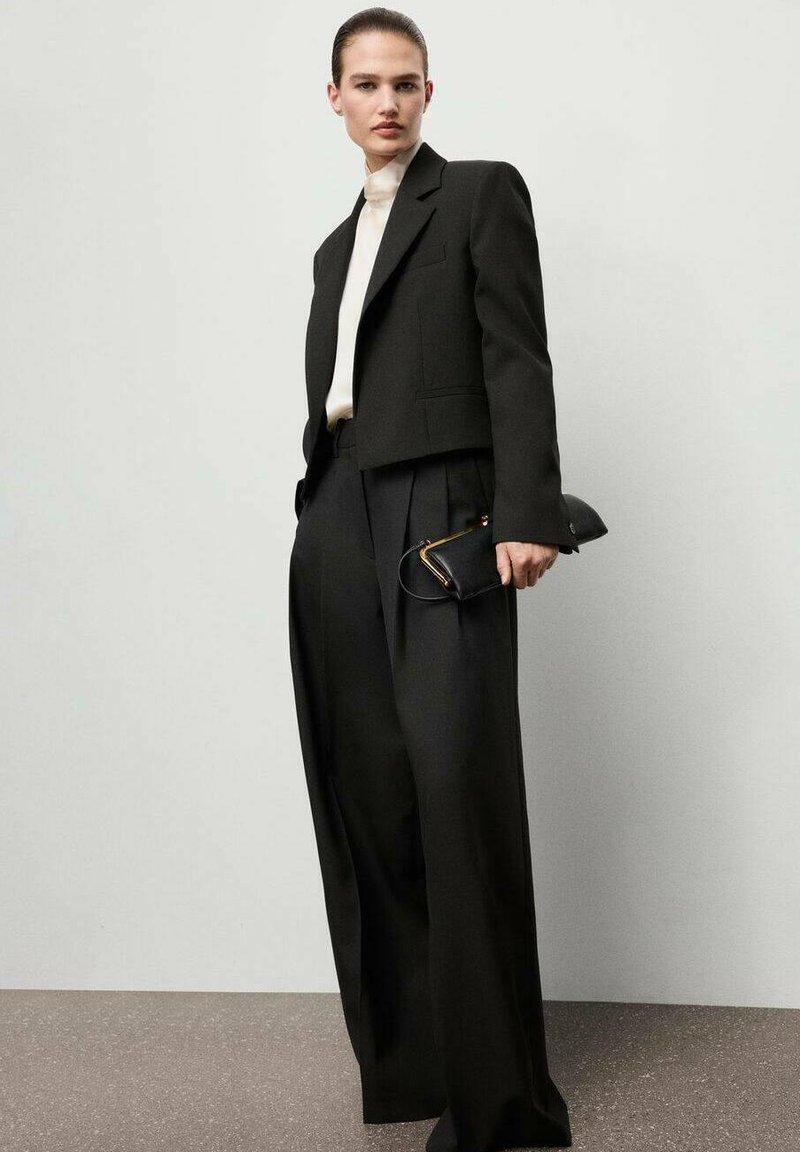 Blazer nero corto su un dolcevita crema, abbinato a pantaloni neri a vita alta e a gamba larga. Tiene una piccola clutch nera.