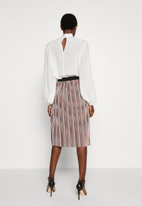 Henkilö, jolla on valkoinen pitkähiha blouse, jossa on avokselta takana, sekä pleatattu midi-hame, jossa on pystysuorat moniväriset raidat, seisoo koroissa.