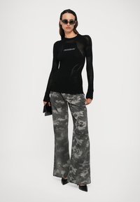 Top negro de manga larga con diseño de malla y logo, combinado con pantalones de camuflaje gris de pierna ancha y tacones negros de punta. Los acentos incluyen gafas de sol.