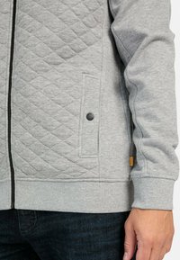 Veste matelassée grise avec fermeture éclair, poignets élastiques et une poche latérale avec un bouton-pression noir. Le tissu est lisse avec une texture douce.