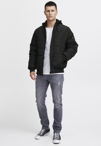 Sort pufferjakke med hætte, der har lynlås og trykknaplukning, båret over en hvid T-shirt og slidte grå jeans. Sorte sneakers fuldender looket.