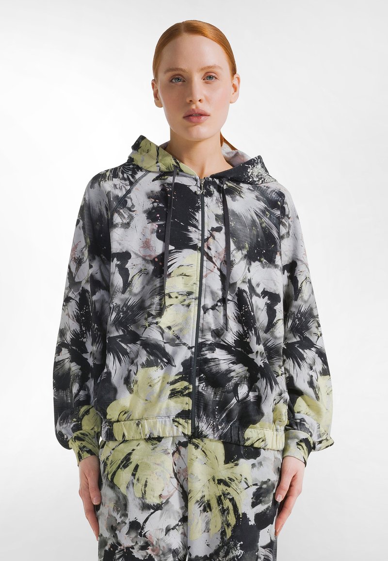 Mujer con cabello rojo vistiendo una sudadera con capucha con cremallera de flores en negro, gris y amarillo pálido, y pantalones a juego, posando contra un fondo blanco.