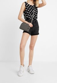 Žena v topu bez rukávů s černými puntíky, černých šortkách s vysokým pasem, bílých teniskách a držící malou šedou crossbody tašku.