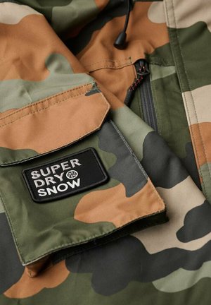 Camo-mønstret stof i grønne og brune nuancer. Har en sort rektangulær lap med "SUPERDRY SNOW" tekst broderet i hvid.