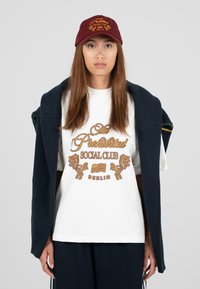 Weißes T-Shirt mit goldfarbigem Grafiktext "Der Verbotene Sozialclub Berlin" und Löwendesigns, kombiniert mit einem marineblauen Cardigan und einer kastanienroten Mütze.