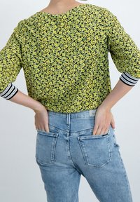 Żółta bluzka w kwiaty z czarnymi akcentami i rękawami za łokieć, zestawiona z jasnoniebieskimi, wysokim stanem dżinsami z tylnymi kieszeniami.