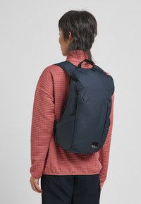 Marineblauwe rugzak met een geborduurde textuur, verstelbare banden en een bovenhandvat, gedragen over een lichtrode gewatteerde jas. Beschikt over zijzakken.