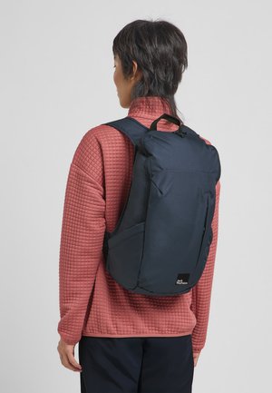 Navy blue backpack with quilted texture, adjustable straps, and top handle, worn over a light red padded jacket. Features side pockets.