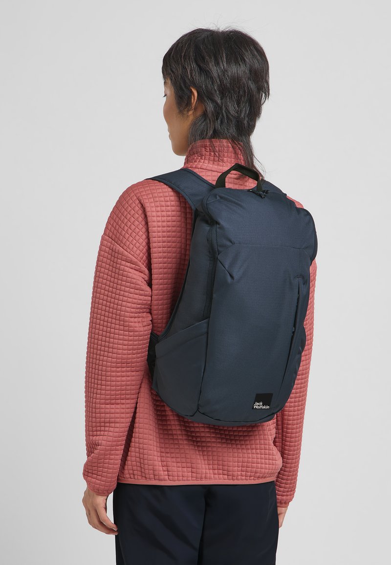 Marineblauwe rugzak met een geborduurde textuur, verstelbare banden en een bovenhandvat, gedragen over een lichtrode gewatteerde jas. Beschikt over zijzakken.