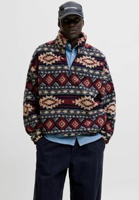 Suéter de forro polar rojo y marino con un diseño estampado y cuello con media cremallera, colocado sobre una camisa azul clara y combinado con pantalones oscuros.
