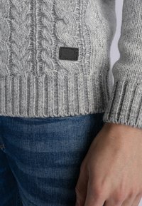 Pulover gri tricotat cu un model de cablu, cu manșete și tiv ribbate, având un eticheta mică de culoare închisă. Purtați cu blugi de denim albastri.