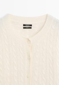 Wool cardigan in cream with a cable knit pattern, featuring a V-neck, five button closures, and labeled with brand and care information.