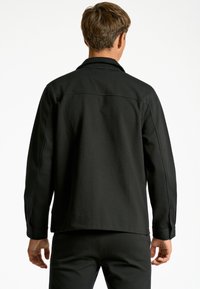 Chaqueta negra con mangas largas, cuello estructurado y puños abotonados. Tejido suave y ligeramente texturizado, con costuras visibles en la parte posterior.