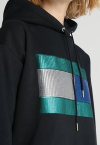 Czarna bluza z kapturem z teksturowaną, kolorową grafiką w teals, srebrze i niebieskich paskach. Zawiera sznurek i metalowe akcenty.