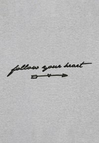 Un tessuto grigio chiaro presenta un testo impresso in nero che recita "segui il tuo cuore", con un cuore e una freccia integrati nel design.