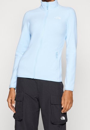 Mujer con chaqueta polar azul claro con cremallera completa y pantalones cargo negros, ambos con el logo visible de The North Face.