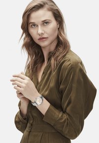 Silberne Uhr mit rundem Zifferblatt, orange- und silberfarbenem Zifferblatt, Netzband. Das Modell trägt eine strukturierte olivgrüne Bluse mit Knopfdetails.