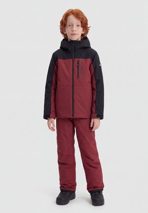 Дете с червена коса, облечено в двуцветно бордо и черно яке и подходящи панталони, стои пред светъл едноцветен фон.
