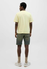 Lysgul bomulds t-shirt, afslappet fit, parret med grønne denim-sorte. Hvide sneakers og beige sokker fuldender outfittet.