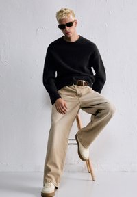 Crni pleteni džemper, bež traperice širokih nogavica, smeđi remen, bijele tenisice i tamne sunčane naočale. Model sjedi na drvenoj stolici ispred teksturiranog zida.