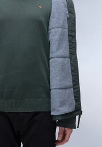 Grøn strikket sweater med en grå polstret ærme og mørkegrønne detaljer. Har et lille flaglogo på brystet og ribbet kant.