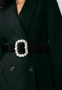 Sort ruskindsbælte med en stor rektangulær spænde udsmykket med perler og klare ædelsten, båret over en mørkegrøn blazer.