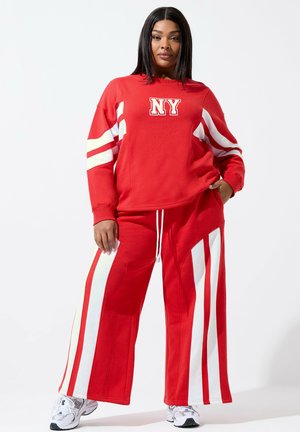 Vrouw draagt een rood trainingspak met witte strepen en "NY" op de trui, staat met één hand in de zak tegen een effen achtergrond.
