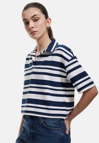 Polo a righe blu navy e bianche con colletto e maniche corte. Il tessuto ha una texture a costine. Indossato con pantaloni shorts in denim a vita alta.
