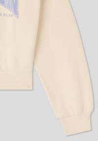 Felpa color crema con polsini a costine, caratterizzata da una vestibilità morbida e una consistenza soffice. Il design include un logo stampato in azzurro chiaro.