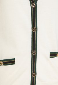 Cardigan blanc avec galon rayé vert et noir, boutons dorés et galon assorti sur deux poches avant.