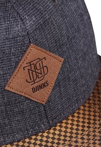 Casquette en tissu sombre avec un dessus texturé gris et une visière à carreaux marron. Un patch en cuir en forme de diamant cousu arbore le logo "DJINNS".