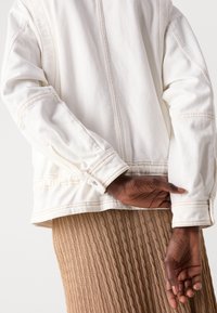 White denim jacket with visible stitching, slightly relaxed fit, and buttoned cuff detail. Worn over a ribbed brown skirt.