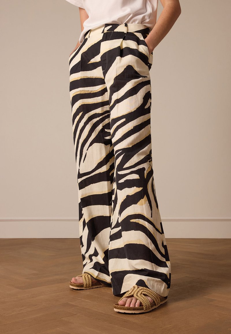 Pantaloni a gamba larga bianchi e neri con un motivo a strisce di zebra, caratterizzati da pieghe e tasche laterali. Indossati con sandali intrecciati di colore neutro.